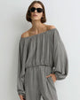 Person modeling a long-sleeve off-the-shoulder jumpsuit with a small-scale black and white pattern, featuring a gathered waistband, one hand in pocket, and paired with sunglasses against a plain light background.