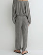 Back view of a person modeling a long-sleeve off-the-shoulder jumpsuit with a small-scale brown and white pattern, featuring a relaxed fit, elastic waistband, and loose straight-leg pants, paired with black flat sandals against a plain light background.