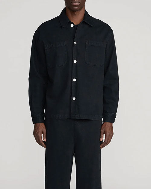 Person modeling a black overshirt-style jacket with a pointed collar, white button front, and two large chest patch pockets, paired with matching black trousers. The outfit is shown from the shoulders to the ankles against a light gray background.