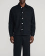 Person modeling a black overshirt-style jacket with a pointed collar, white button front, and two large chest patch pockets, paired with matching black trousers. The outfit is shown from the shoulders to the ankles against a light gray background.