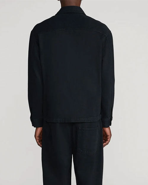 Back view of a model wearing a black long-sleeve overshirt with a pointed collar, straight hem, and back yoke seam, paired with matching black trousers featuring a large patch pocket on the back. Shown from the shoulders to below the knees against a light gray background.