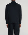 Back view of a model wearing a black long-sleeve overshirt with a pointed collar, straight hem, and back yoke seam, paired with matching black trousers featuring a large patch pocket on the back. Shown from the shoulders to below the knees against a light gray background.
