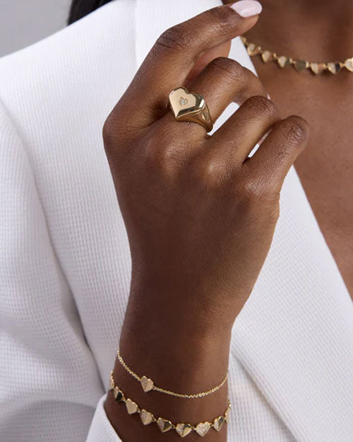 Close-up of a hand wearing gold jewelry: a heart-shaped ring with an engraved detail on the middle finger, a gold bracelet with heart-shaped charms, and a simple chain bracelet, paired with a white textured garment.
