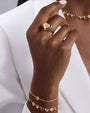 Close-up of a hand wearing gold jewelry: a heart-shaped ring with an engraved detail on the middle finger, a gold bracelet with heart-shaped charms, and a simple chain bracelet, paired with a white textured garment.