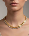 Close-up of a person wearing a layered necklace featuring two rows: one with large gold curb chain links and another with sparkling round-cut crystals, resting on the collarbone and paired with a light V-neck top.