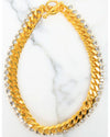 A gold-toned necklace featuring two layers: one with large curb chain links and another with a row of clear round-cut crystals, displayed on a white marble surface.