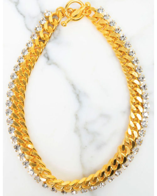 A gold-toned necklace featuring two layers: one with large curb chain links and another with a row of clear round-cut crystals, displayed on a white marble surface.