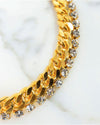 Close-up of a gold-toned curb chain necklace paired with a row of clear round-cut crystals, displayed on a white marble surface.