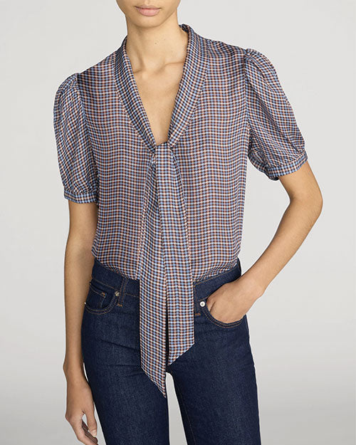 Front view of a lightweight small-check print blouse in blue, brown, and cream tones. The blouse features a deep V-neckline with a self-tie scarf detail at the collar, short puff sleeves, and a relaxed fit. The blouse is tucked into dark indigo jeans with classic five-pocket styling. One hand rests in a front pocket. The image is shown against a light gray background.