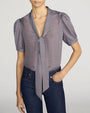 Front view of a lightweight small-check print blouse in blue, brown, and cream tones. The blouse features a deep V-neckline with a self-tie scarf detail at the collar, short puff sleeves, and a relaxed fit. The blouse is tucked into dark indigo jeans with classic five-pocket styling. One hand rests in a front pocket. The image is shown against a light gray background.