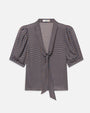 Front view of a short-sleeve blouse featuring a small blue, brown, and cream check pattern. The blouse has a deep V-neckline with a self-tie scarf detail at the front, gathered shoulders, and softly puffed sleeves ending just above the elbows. The lightweight fabric drapes loosely through the body, and the blouse is displayed against a light gray background.