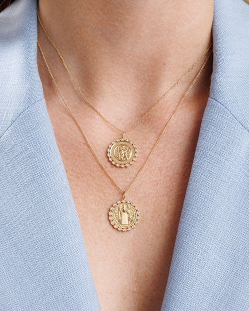 Close-up of a person wearing two layered gold necklaces with round medallion pendants. Each pendant has a scalloped edge adorned with small gemstones. The top pendant features an engraved Saint Christopher design, while the bottom pendant shows a different religious figure in the center.