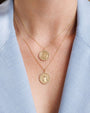 Close-up of a person wearing two layered gold necklaces with round medallion pendants. Each pendant has a scalloped edge adorned with small gemstones. The top pendant features an engraved Saint Christopher design, while the bottom pendant shows a different religious figure in the center.