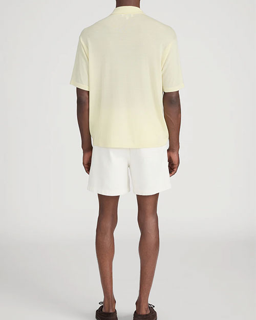 Back view of a person standing in a studio against a light neutral background, wearing a pale yellow short-sleeve polo shirt with a soft collar and button placket, paired with off-white drawstring shorts. 
