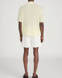 Back view of a person standing in a studio against a light neutral background, wearing a pale yellow short-sleeve polo shirt with a soft collar and button placket, paired with off-white drawstring shorts. 