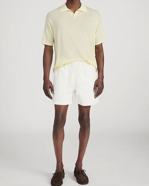 Person standing in a studio against a light neutral background, wearing a pale yellow short-sleeve polo shirt with a soft collar and button placket, paired with off-white drawstring shorts. The outfit is styled with brown lace-up shoes, and the image is photographed in a full-length, front-facing pose.