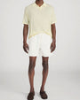 Person standing in a studio against a light neutral background, wearing a pale yellow short-sleeve polo shirt with a soft collar and button placket, paired with off-white drawstring shorts. The outfit is styled with brown lace-up shoes, and the image is photographed in a full-length, front-facing pose.