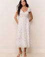 White ankle-length dress with short puff sleeves, lace details, and subtle floral pattern, paired with strappy heels.
