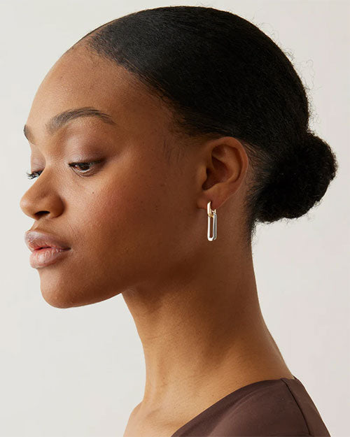 Close-up of an ear wearing a small, modern gold hoop earring with a rectangular drop design, hair styled in a neat low bun.