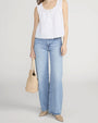 Person standing in a studio against a light neutral background, wearing a white sleeveless blouse with scalloped edges along the neckline and hem, featuring a button-front design. The blouse is paired with mid-rise, straight-leg light-wash jeans and pointed-toe shoes. The look is accessorized with a woven straw tote bag, photographed in a full-length, front-facing pose.