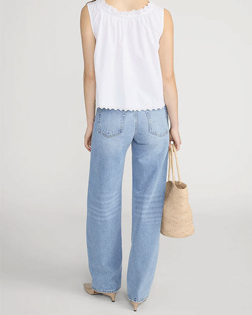 Rear-view studio image of a person standing against a light neutral background, wearing a white sleeveless blouse with scalloped edges along the neckline and hem. The blouse is paired with mid-rise, straight-leg light-wash jeans featuring back patch pockets and subtle fading. A woven straw tote bag is held at the side, and pointed-toe shoes are visible, photographed in a full-length, back-facing pose.