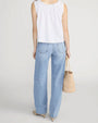 Rear-view studio image of a person standing against a light neutral background, wearing a white sleeveless blouse with scalloped edges along the neckline and hem. The blouse is paired with mid-rise, straight-leg light-wash jeans featuring back patch pockets and subtle fading. A woven straw tote bag is held at the side, and pointed-toe shoes are visible, photographed in a full-length, back-facing pose.