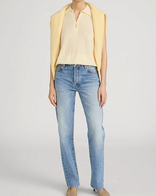Person standing in a studio against a light neutral background, wearing a soft yellow sleeveless knit polo-style top with a short zipper neckline. A matching yellow sweater is draped over the shoulders. The outfit is paired with mid-rise, straight-leg light-wash jeans and neutral-toned shoes, photographed in a full-length, front-facing pose.