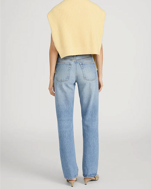 Rear-view studio image of a person standing against a light neutral background, wearing a pale yellow ribbed sweater draped over the shoulders like a shawl. The outfit is paired with mid-rise, straight-leg light-wash jeans featuring back patch pockets and subtle fading. Neutral-toned heeled shoes are visible, and the image is photographed in a full-length, back-facing pose.