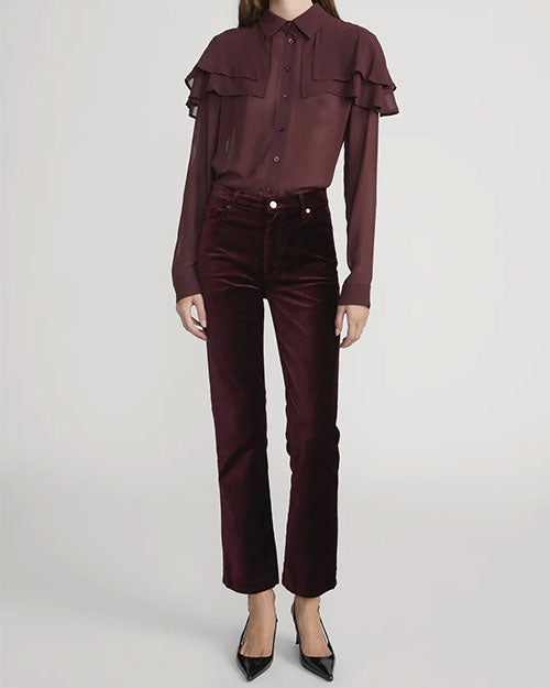 Person wearing a long-sleeve button-up blouse with layered ruffle details in a deep burgundy color, paired with high-waisted velvet pants in a matching shade and pointed black shoes.
