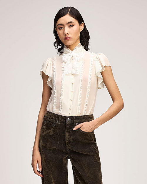 Ivory ruffle-trim blouse with a high neck tie, flutter sleeves, and front button detailing, paired with dark washed jeans.