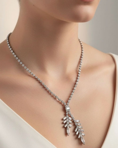 A close-up of a person wearing a silver-toned necklace with a Y-shaped design, featuring two dangling branch-like pendants made of stacked metallic bars. The necklace rests on the collarbone and is paired with a light V-neck top.