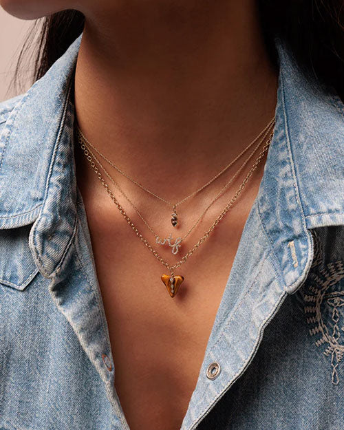 Close-up of a neck wearing three layered necklaces: a thin chain with a small pendant, a middle chain with “wtf” letters, and a bottom chain with a triangular pendant featuring an intricate design. The person is wearing a denim shirt.