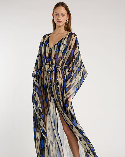 Person standing in a studio against a light neutral background, wearing a sheer, floor-length patterned cover-up with a deep V-neckline and flowy sleeves. The fabric features geometric shapes in shades of brown, blue, beige, and black. A coordinating bikini top is visible underneath, and the look is styled with strappy heeled sandals, photographed in a full-length, front-facing pose.