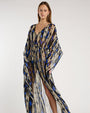 Person standing in a studio against a light neutral background, wearing a sheer, floor-length patterned cover-up with a deep V-neckline and flowy sleeves. The fabric features geometric shapes in shades of brown, blue, beige, and black. A coordinating bikini top is visible underneath, and the look is styled with strappy heeled sandals, photographed in a full-length, front-facing pose.