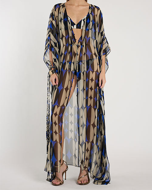 Person standing in a studio against a light neutral background, wearing a sheer, floor-length patterned cover-up with a deep V-neckline and flowy sleeves. The fabric features geometric shapes in shades of brown, blue, beige, and black. A coordinating bikini top is visible underneath, and the look is styled with strappy heeled sandals, photographed in a full-length, front-facing pose.