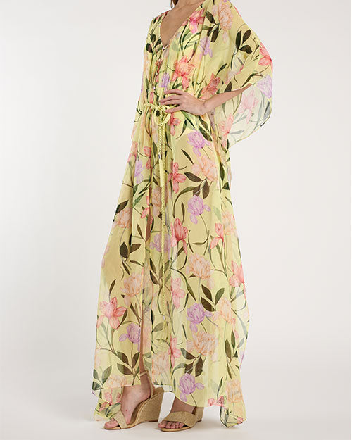 Person standing in a studio against a light neutral background, wearing a sheer, floor-length yellow cover-up with a V-neckline, flowing sleeves, and an adjustable waist tie. The fabric features a floral pattern in shades of pink, green, and lavender. Open-toe heeled sandals are visible, and the image is photographed in a full-length, front-facing pose.