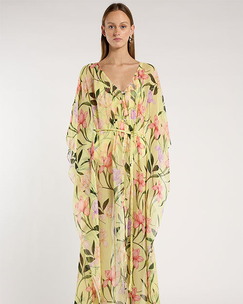 Person standing in a studio against a light neutral background, wearing a sheer, floor-length yellow cover-up with a V-neckline, flowing sleeves, and an adjustable waist tie. The fabric features a floral pattern in shades of pink, green, and lavender. Open-toe heeled sandals are visible, and the image is photographed in a full-length, front-facing pose.