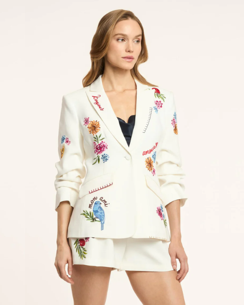 Woman wearing a white embroidered blazer with floral and bird designs on a plain background