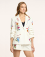 Woman wearing a white embroidered blazer with floral and bird designs on a plain background