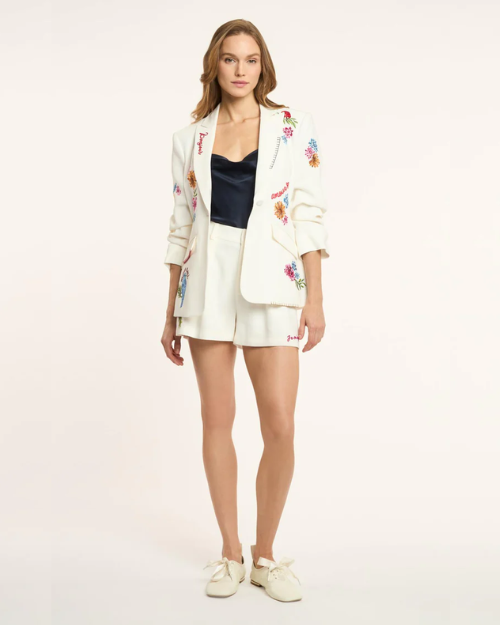 Woman wearing a white embroidered blazer over a black top and white shorts on a light background