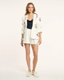 Woman wearing a white embroidered blazer over a black top and white shorts on a light background