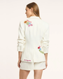 Woman wearing a white blazer with floral embroidery on a plain background