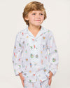 Child wearing a white pajama set featuring a long-sleeved, collared button-up shirt and matching pants, both adorned with colorful illustrations of animals, people, and nature scenes, standing against a plain background.