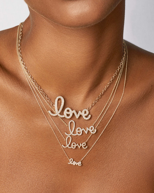 Person wearing multiple layered gold necklaces, each featuring a script-style “love” pendant with small sparkling stones against a white background.