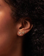 Close-up of a person’s ear adorned with two earrings. The upper earring is shaped like a gold camera, and the lower is a gold pave diamond plane.