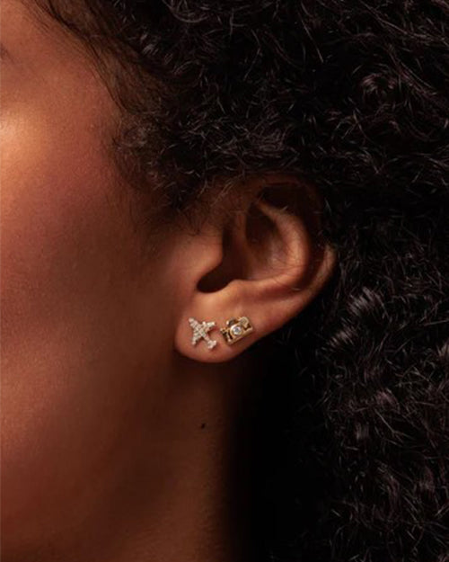 Close-up of a person’s ear adorned with two earrings. The upper earring is shaped like a gold camera, and the lower is a gold pave diamond plane.