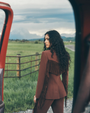Woman in a brown fringe jacket standing in a field with a red vehicle and mountains in the background