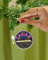 A hand displays a small Christmas ornament inscribed with the word "caviar," adding a unique touch to holiday decor.