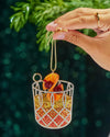 A hand presents a festive Christmas ornament featuring a cocktail served in a glass, with a orange and cherry garnish on top. 