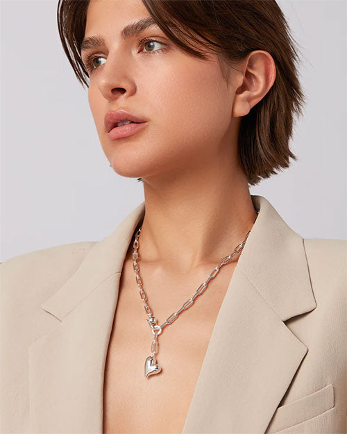 A woman in a white blazer wearing a heart-shaped necklace, smiling confidently at the camera.